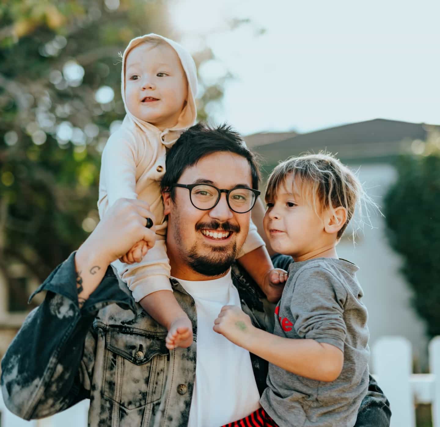 An adult with glasses smiles while holding a young child. A baby sits on top of the adult's shoulders.